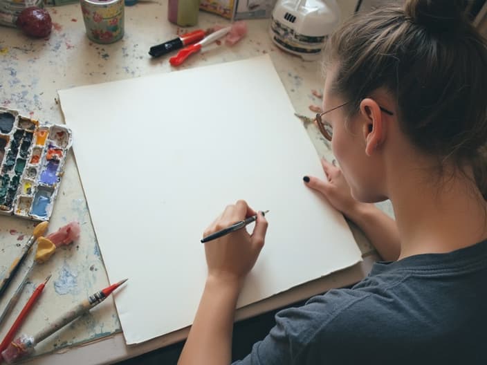 Person looking frustrated at a blank canvas or notebook, surrounded by art supplies